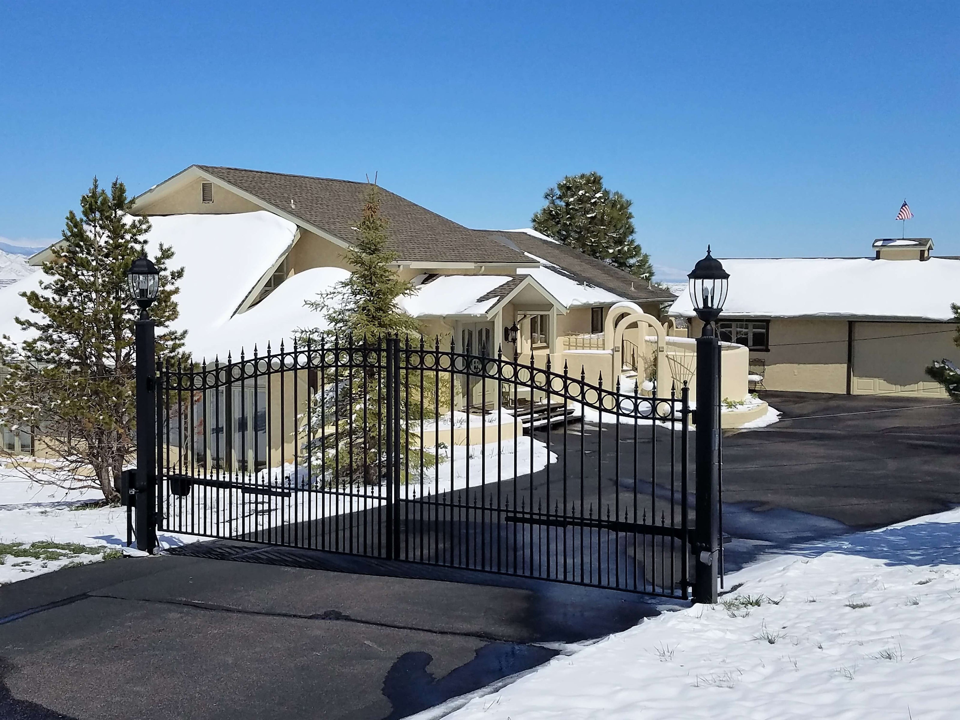 double black iron gate with arched top and row of circles and spear point finials between black metal lampposts on driveway in snow covered yard of tan adobe home