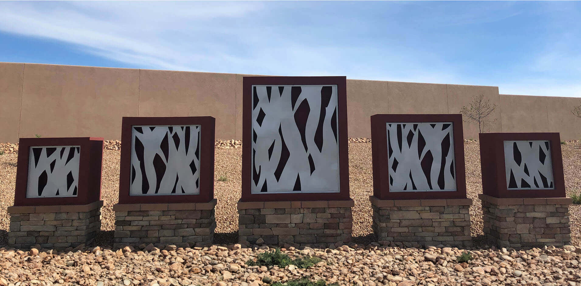 Row of five kiosks mounted on stacked stone with lazer cut stainless steel designs in rust color metal frames in front of adobe wall of neighborhood entrance