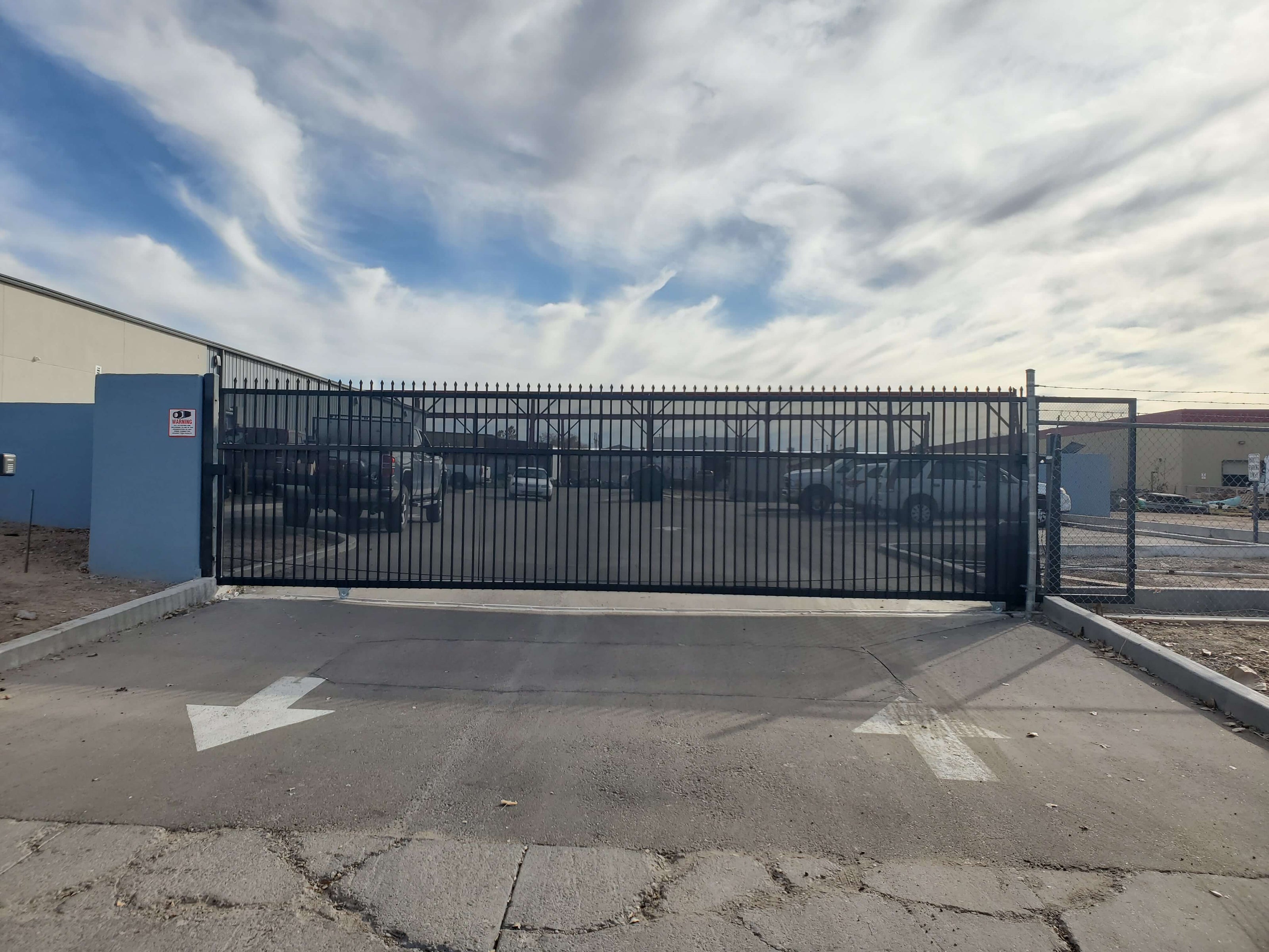 a large sliding commercial gate with vertical bars and privacy screen on an entrance to a warehouse parking lot with cars in background