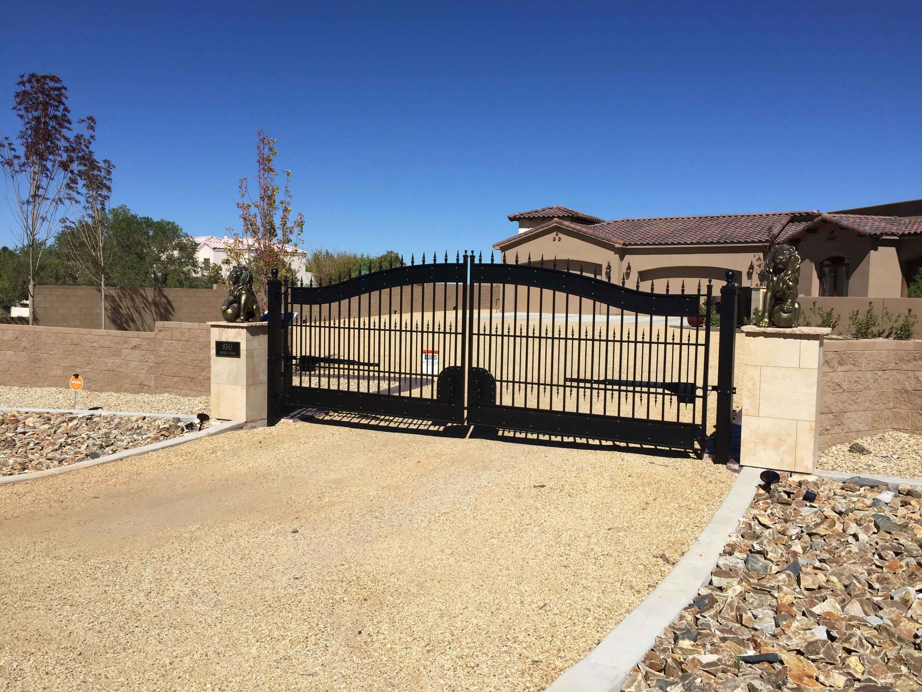 Black double wrought iron gate with heavy solid bands top and botton. Arched with rivets on bands. Gate is on gravel driveway between sandstone pillars. in a concrete block wall.