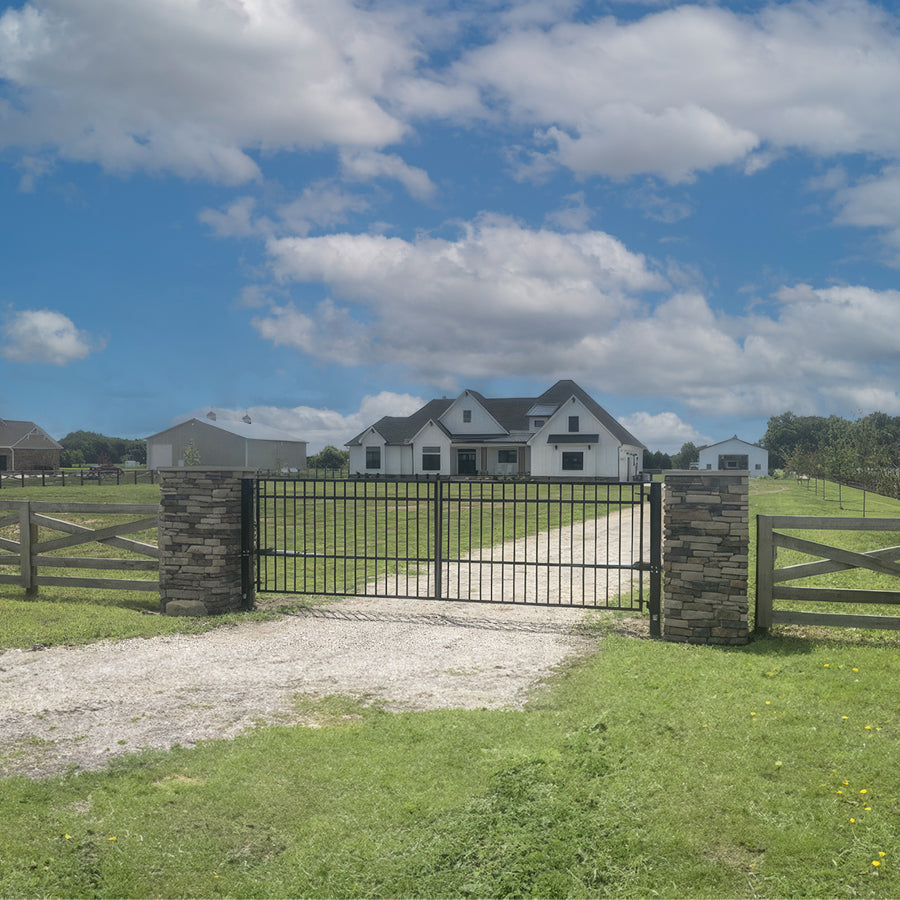 In a green field, under a blue sky, a rectangular double black wrought iron driveway gate sits on a gravel drive leading to large farm house. The simple gate has vertical bars and one horizontal bar near the top and one about a third way from bottom. It is attacked to two stone square pillars in a ranch style wood fence.