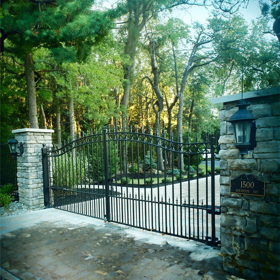 two square stone columns at end of wide driveway with double black wrought iron gate that is arched with row of circles trimming the top and spear point finials