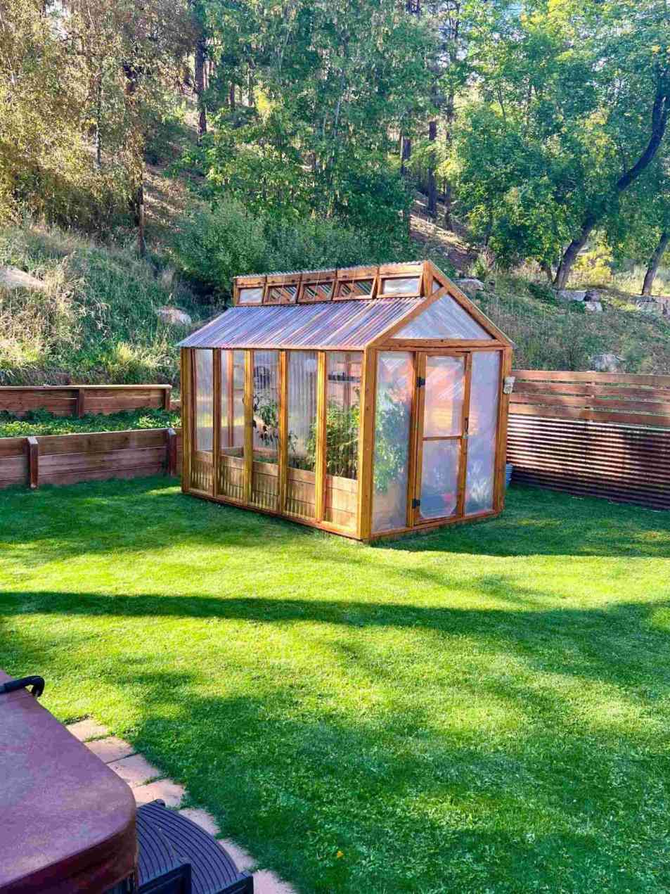 A charming wooden greenhouse with transparent panels sits on a well-manicured lawn, surrounded by lush greenery and trees.