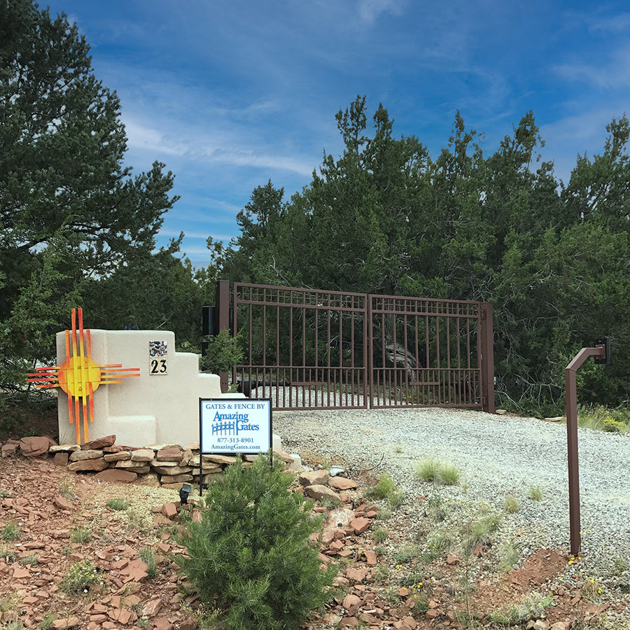 Rust Color double gate on a gravel road bordered by evergreens and red rocks with adobe aztec style mailbox and rust color keypad to open gate