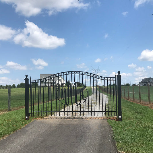 single black wrought iron gate with the  monomgram MMA in an oval in the middle in front of long road on flat green field of grass, big farmhouse in background and blue sky with clouds