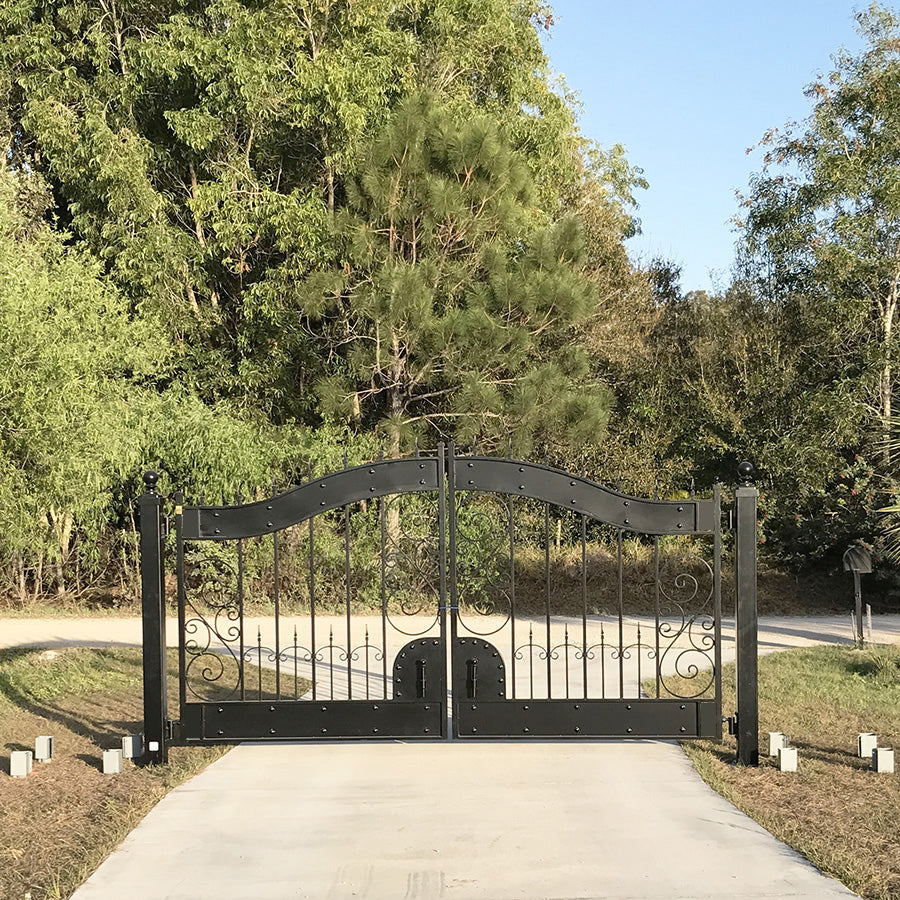 Hyde Park Style ornate arched black wrought iron double driveway gate at the end of a drive on a Florida lawn, palm trees and woods