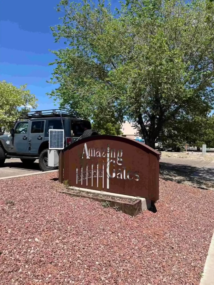 Rust-colored metal sign with cutout lettering and gate design, set on red gravel and parked gray SUV under a blue sky.