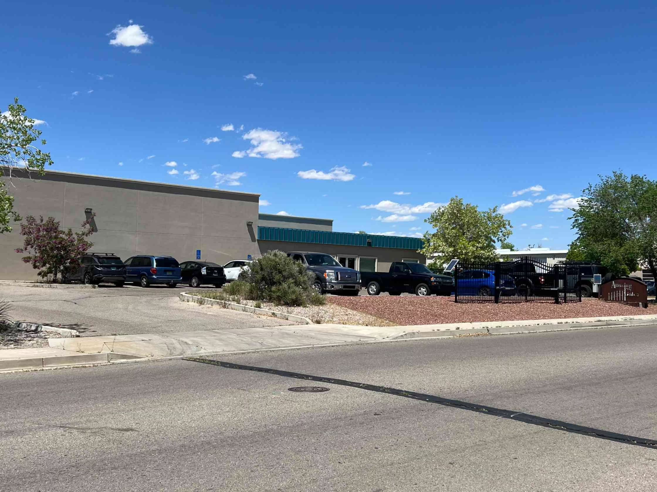 Building with a teal awning, parking lot with vehicles, black metal gate, red gravel landscaping, and clear blue sky.