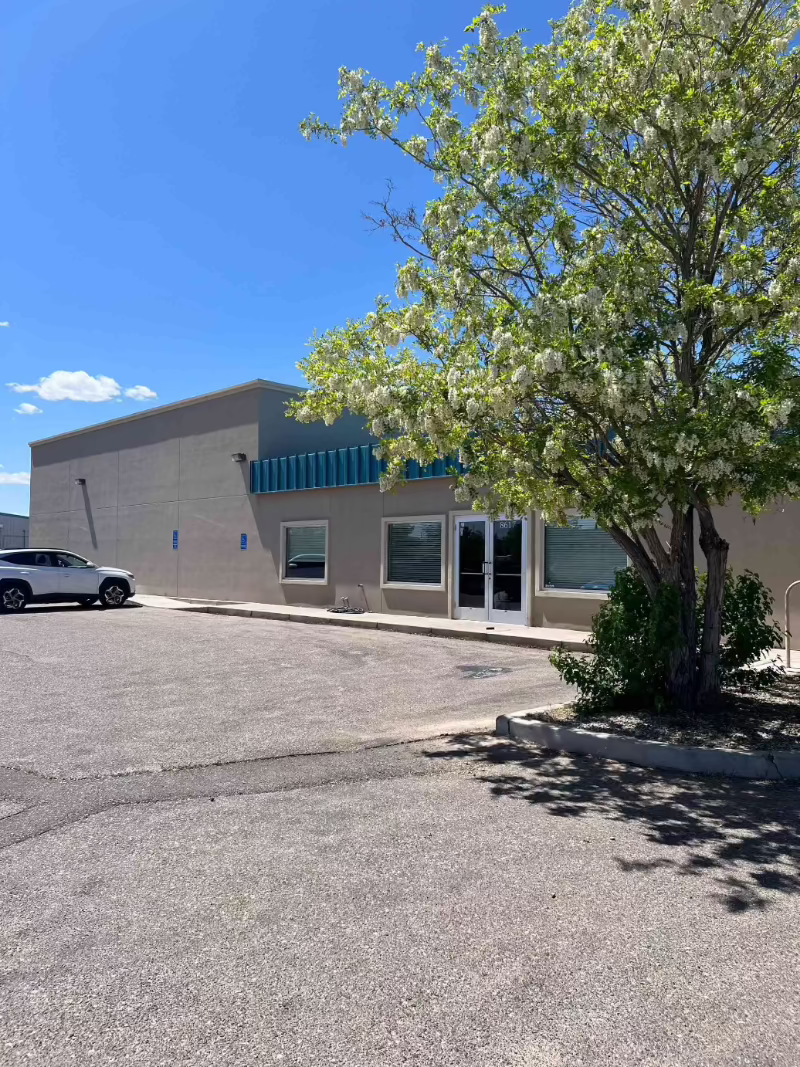 Single-story beige building with teal awning, glass doors, parking lot, and a leafy tree in bloom under a bright blue sky.
