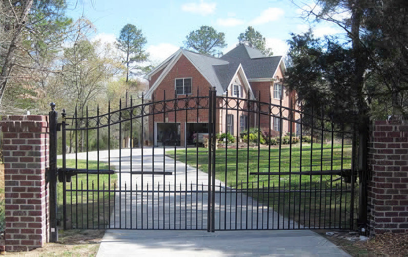 Black wrought iron driveway gate with a curved top, set between brick pillars, leading to a red brick house.