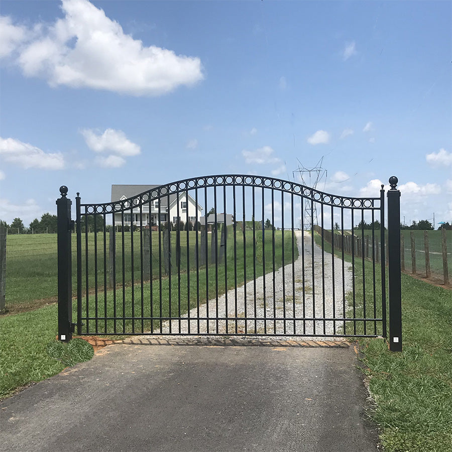 single black wrought iron gate with ball caps on both posts in front of long road on flat green field of grass, big farmhouse in background and blue sky with clouds