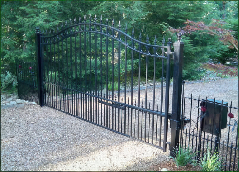 Black wrought iron gate with a curved top and spear-shaped finials, installed on a gravel driveway surrounded by greenery.