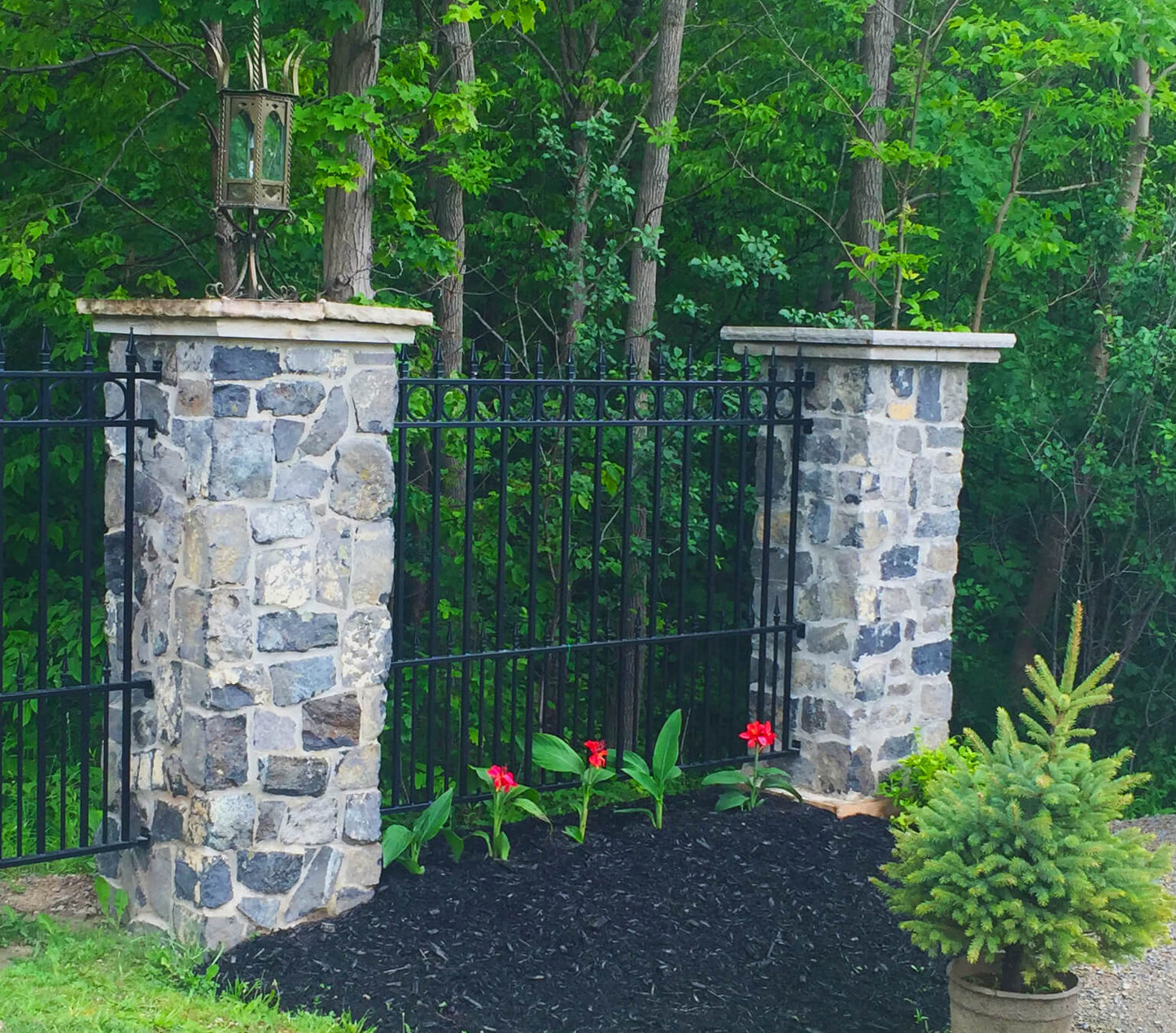 Wrought iron fence installed between stone pillars with decorative lanterns on top, surrounded by vibrant greenery and flowers.