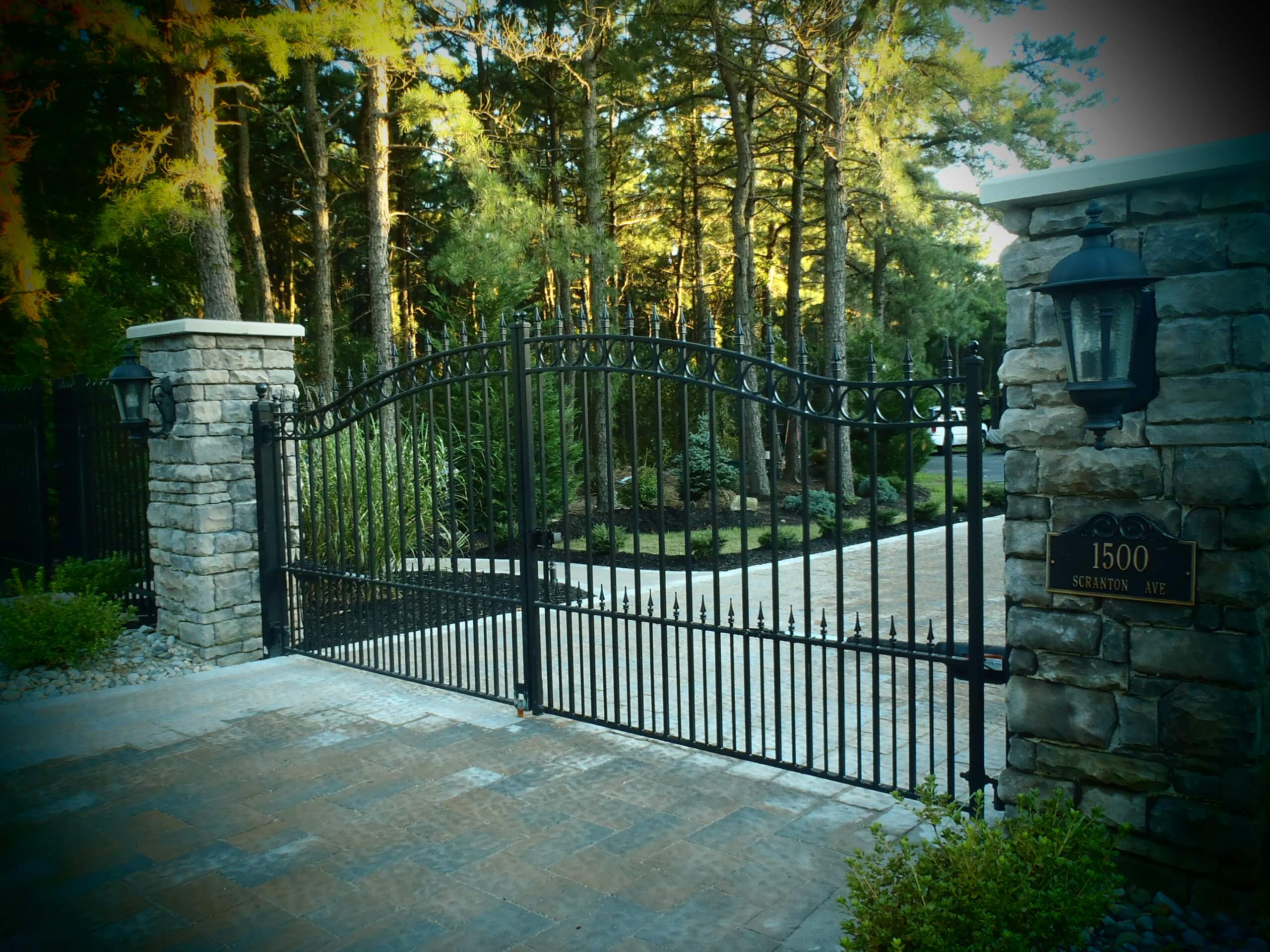 two square stone columns at end of wide driveway with double black wrought iron gate that is arched with row of circles trimming the top and spear point finials
