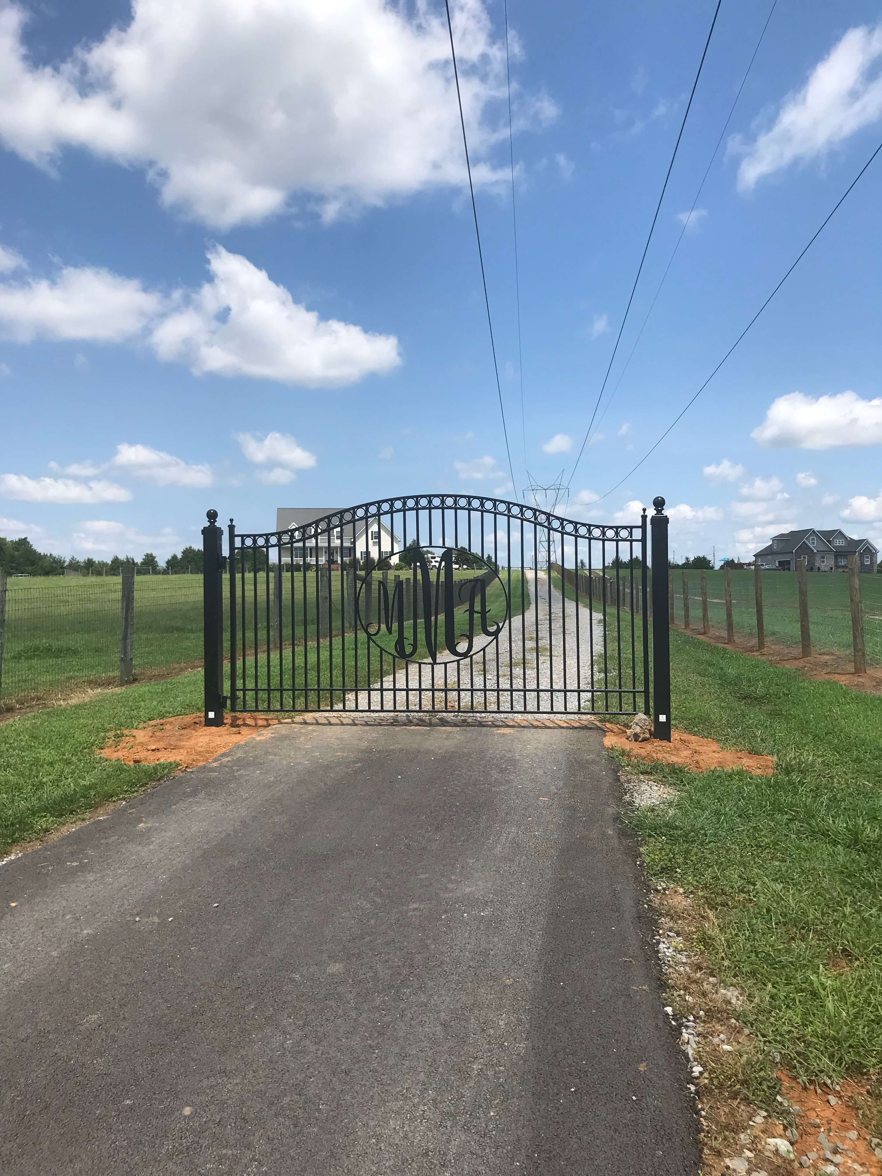 single black wrought iron gate with the  monomgram MMA in an oval in the middle in front of long road on flat green field of grass, big farmhouse in background and blue sky with clouds
