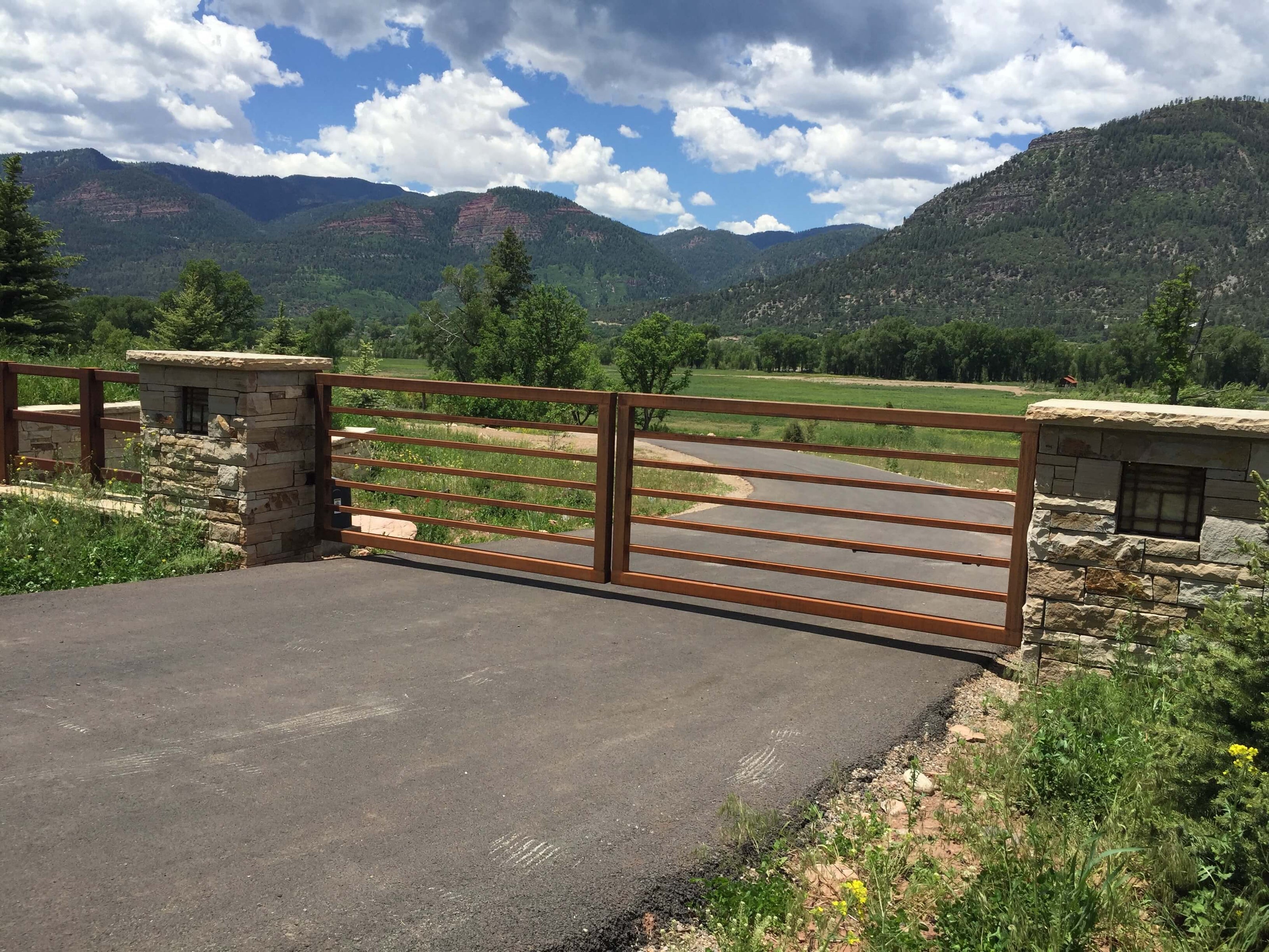 rust color metal double simple gate with horizontal bars in rectangular frame hinged to square stacked stone columns with inset craftsman style lights on paved road in green vallley, red rock mountains in background