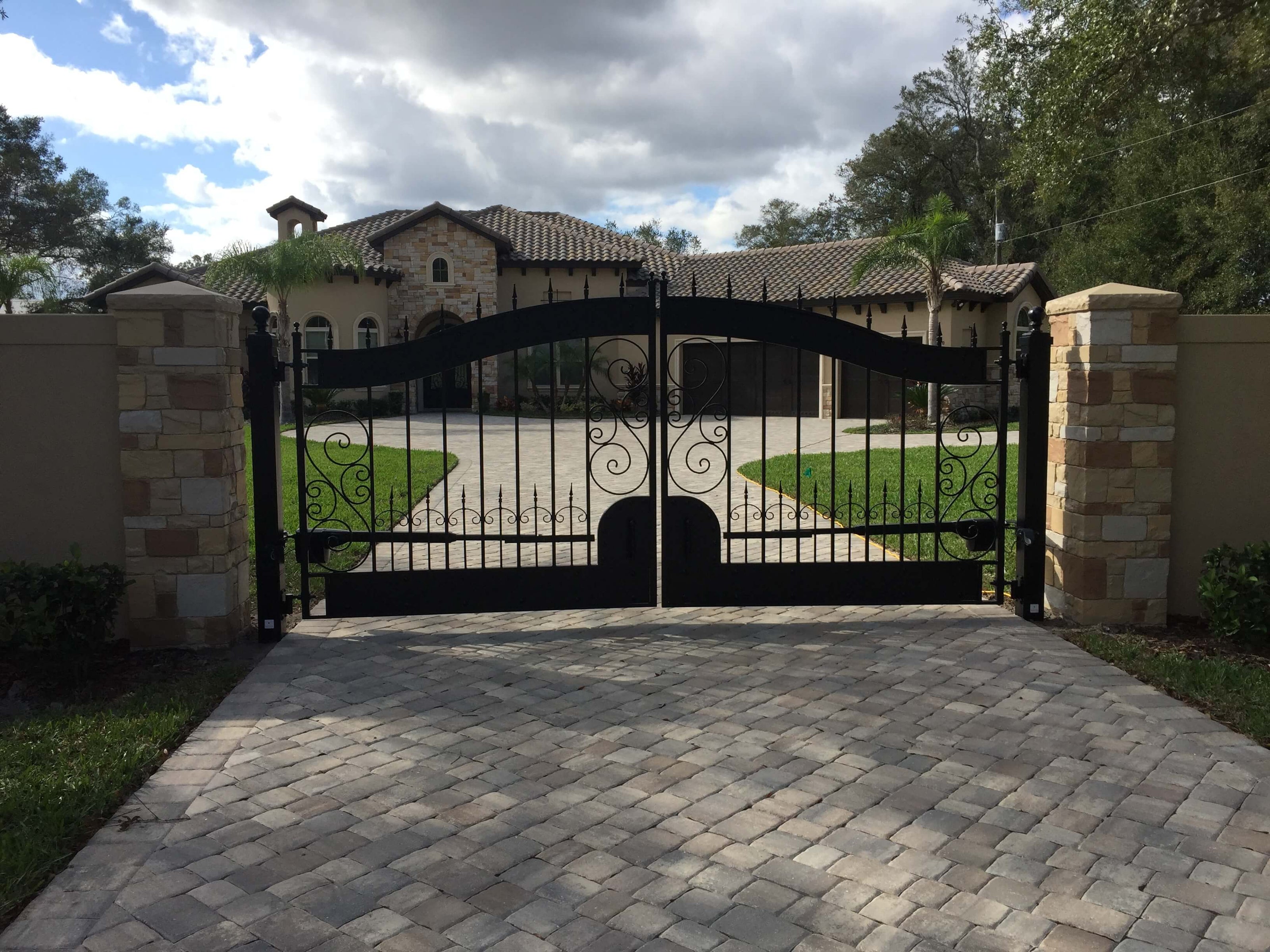 double fancy wrought iron gate with arch, spiral motif set in vertical bars wide bands top and bottom and spear point finials in stone wall on cobblestone driveway in front of large tile roof home