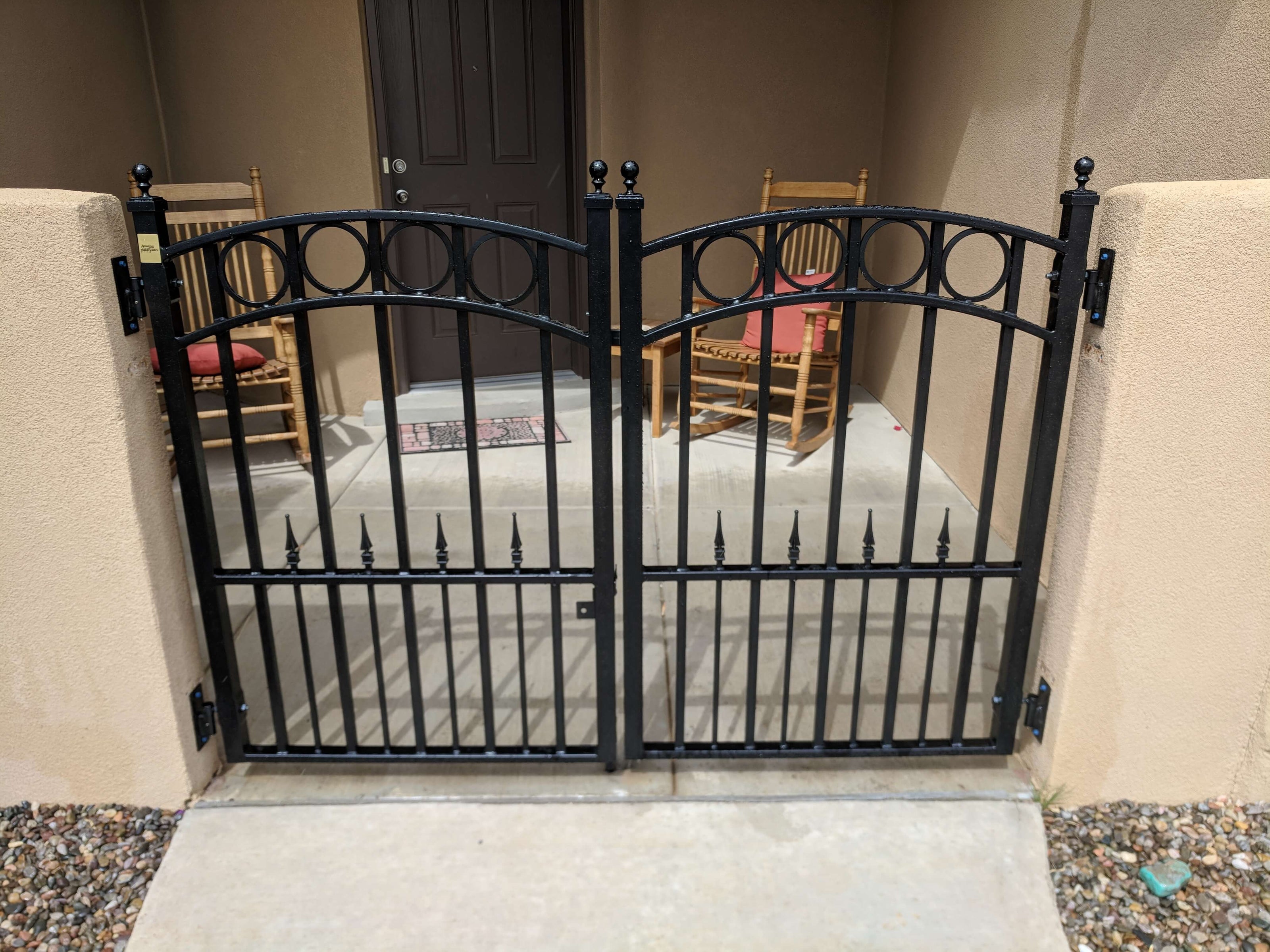 double pedestrian gate in black wrought iron with arch on each gate and row of circles on top, hinged to adobe wall and adobe front porch behind