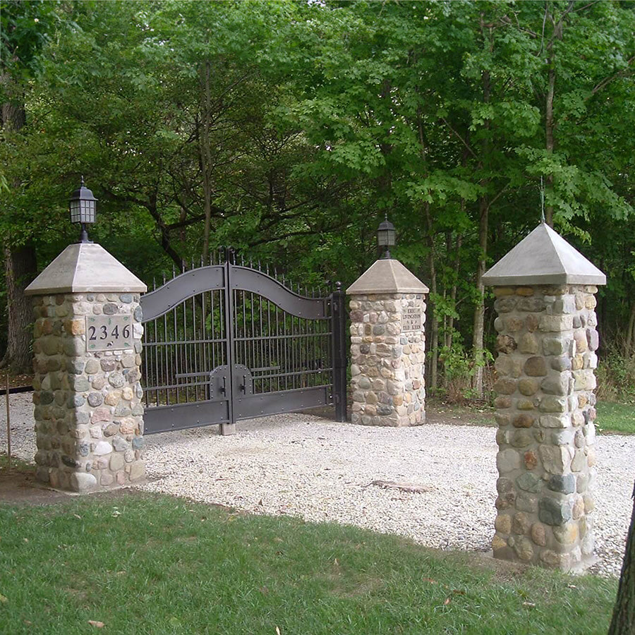 stately double bronze color gate with arch and spear points on top between two square river rock pillars topped with concrete spires holding black lanterns on a gravel driveway in the woods