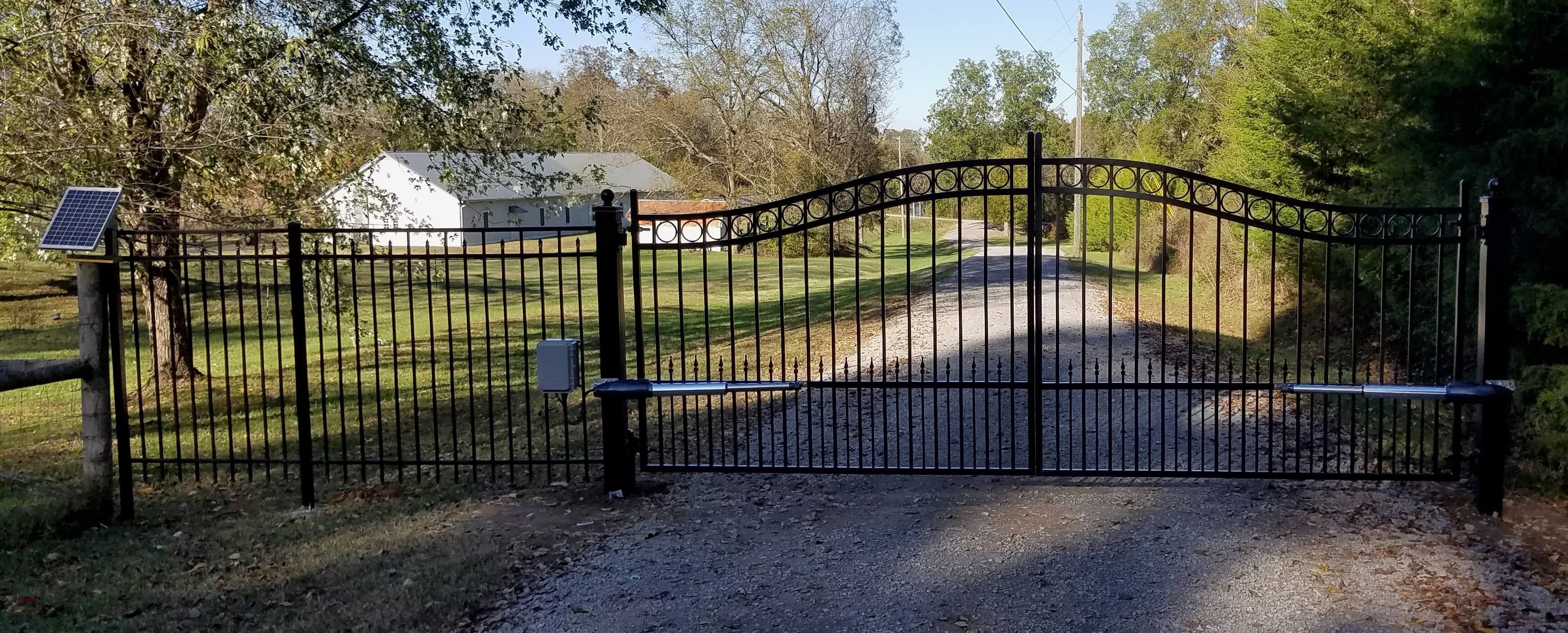double arched black wrought iron gate with row of circles on top ball caps on both posts in front of long gravel driveway with blue sky with clouds
