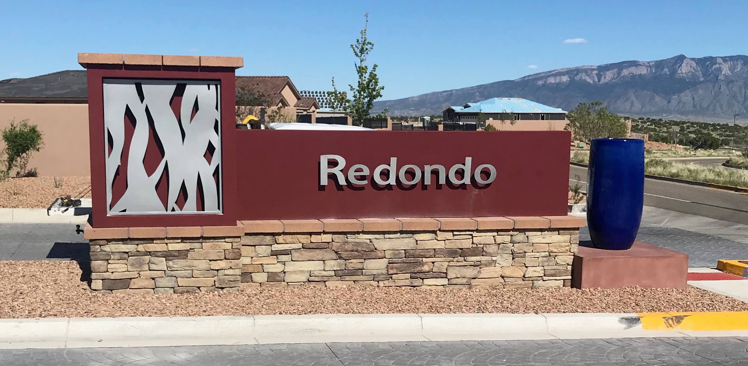 rust color metal sign that says Redondo and stainless steel metal cutout of abstract tree trunks built on stack stone pediment in front of entrance to neighborhood, mountains in backround, southwestern setting