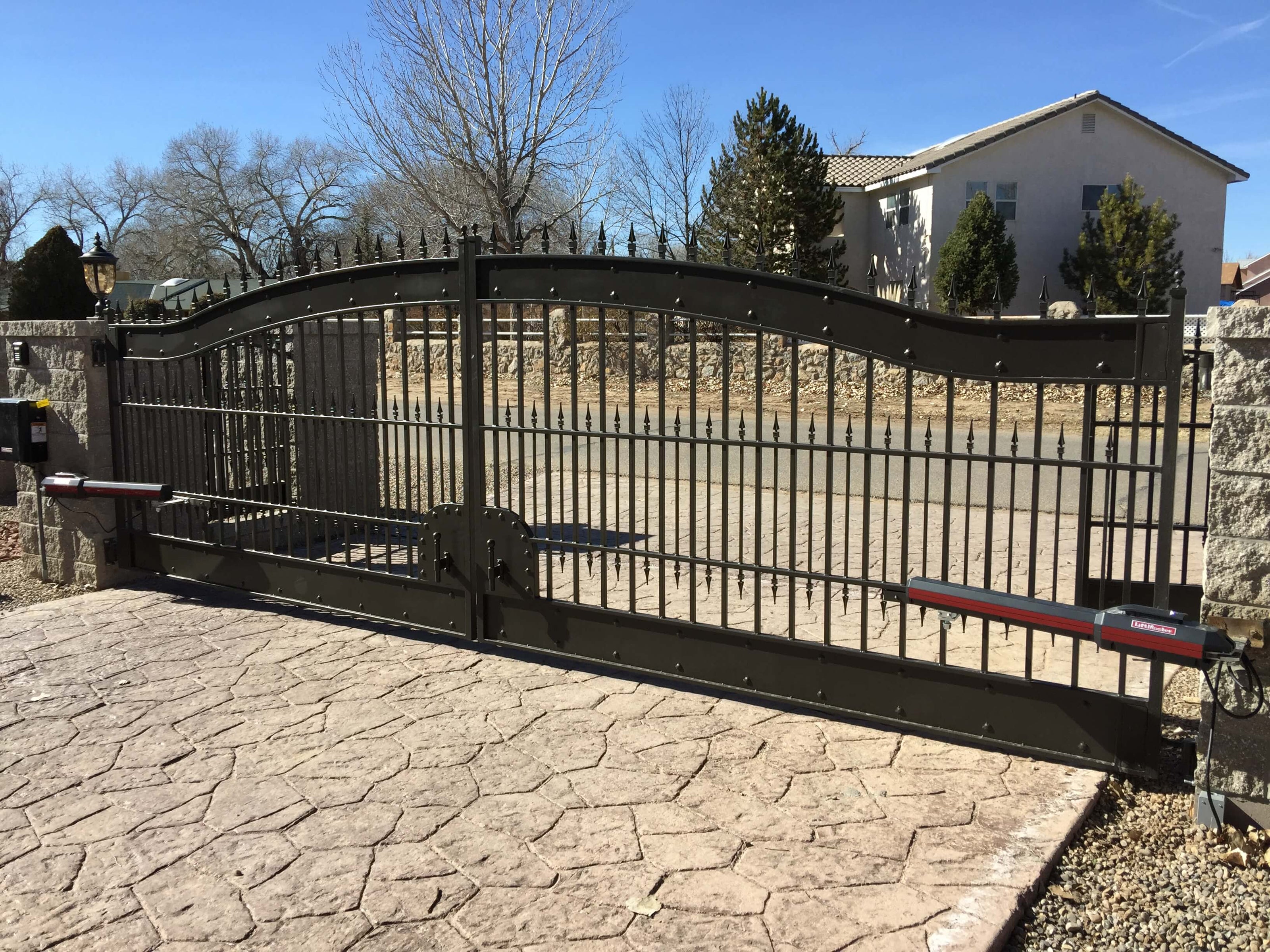 wide bronze color double wrought iron gate with heavy solid bands top and botton. Arched with rivets on bands. Gate is on flagstone driveway mounted between two square stone pillars.