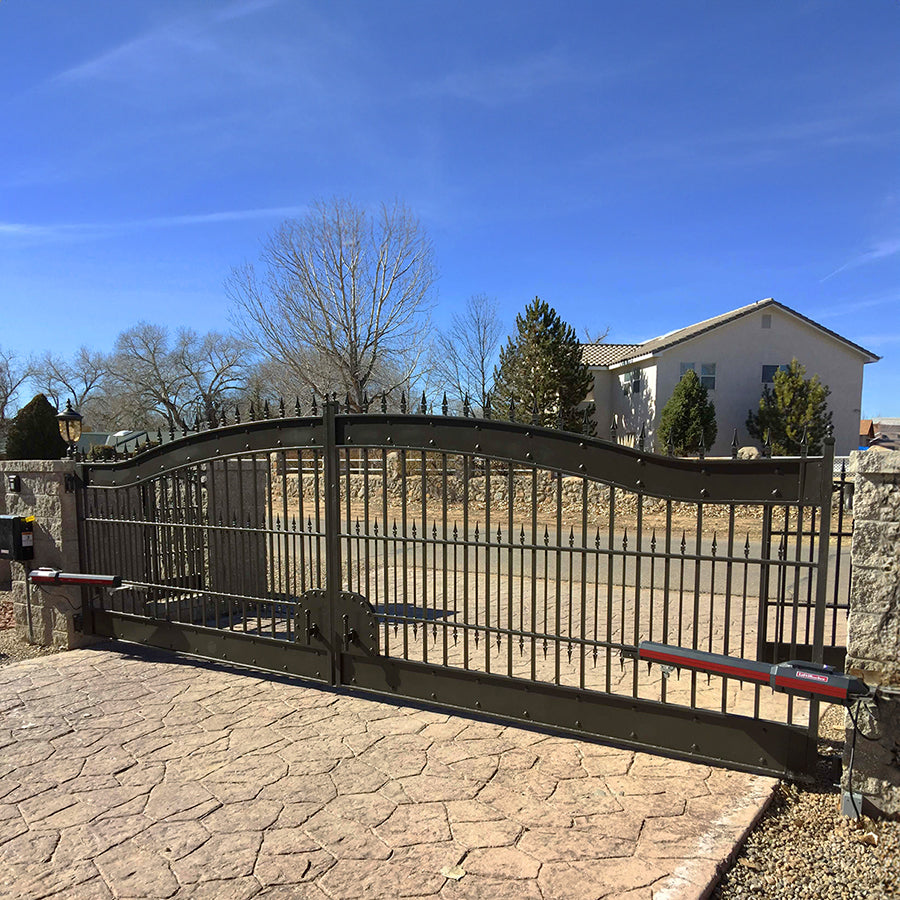 wide bronze color double wrought iron gate with heavy solid bands top and botton. Arched with rivets on bands. Gate is on flagstone driveway mounted between two square stone pillars.