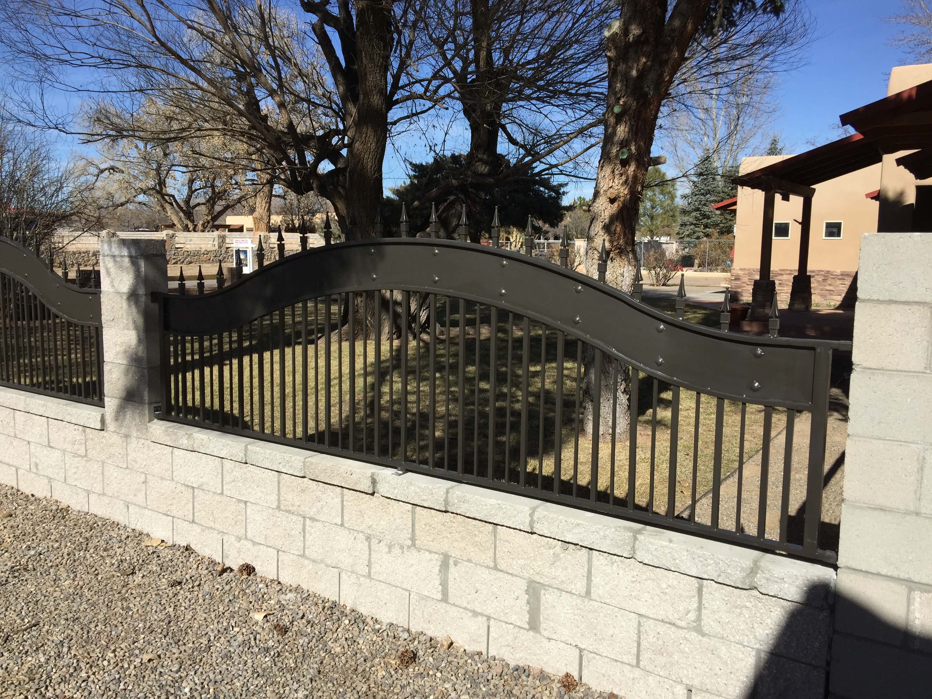 section of bronze color iron fence with arched top wide band trim and spear point finials set in stone block wall around the yard of an adobe