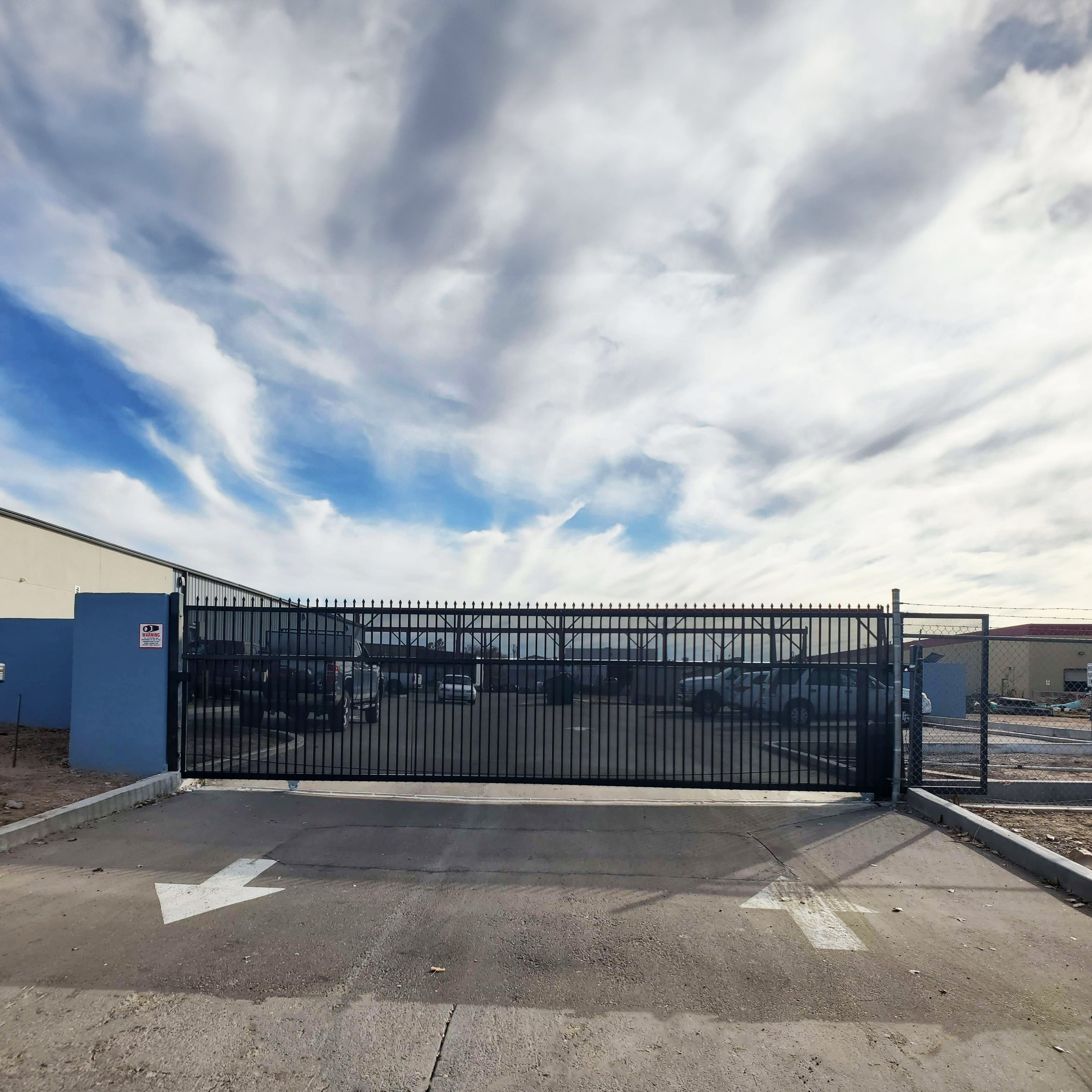 a large sliding commercial gate with vertical bars and privacy screen on an entrance to a warehouse parking lot with cars in background