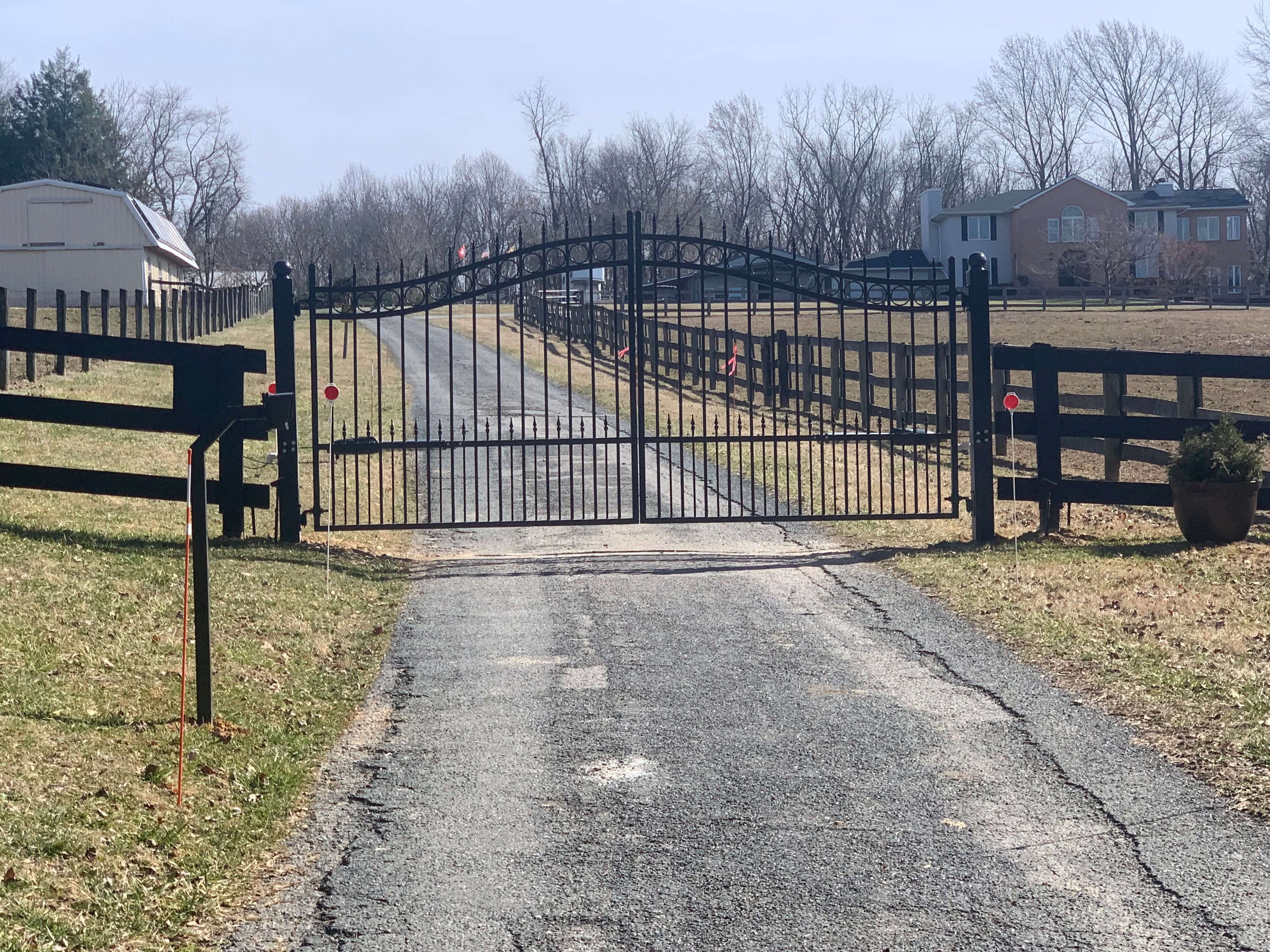 double arched black wrought iron gate with row of circles on top ball caps on both posts in front of long paved driveway and barn in background