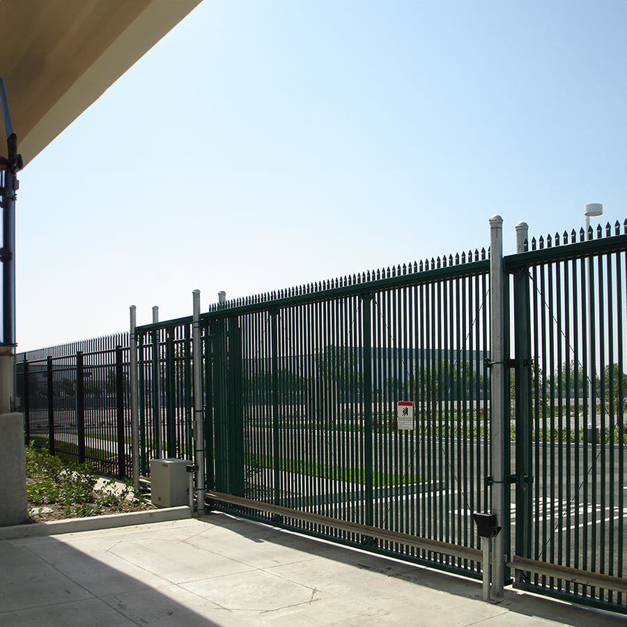 Industrial Black sliding metal security gate with vertical bars & pointed finials enclosing a parking lot under a clear sky.