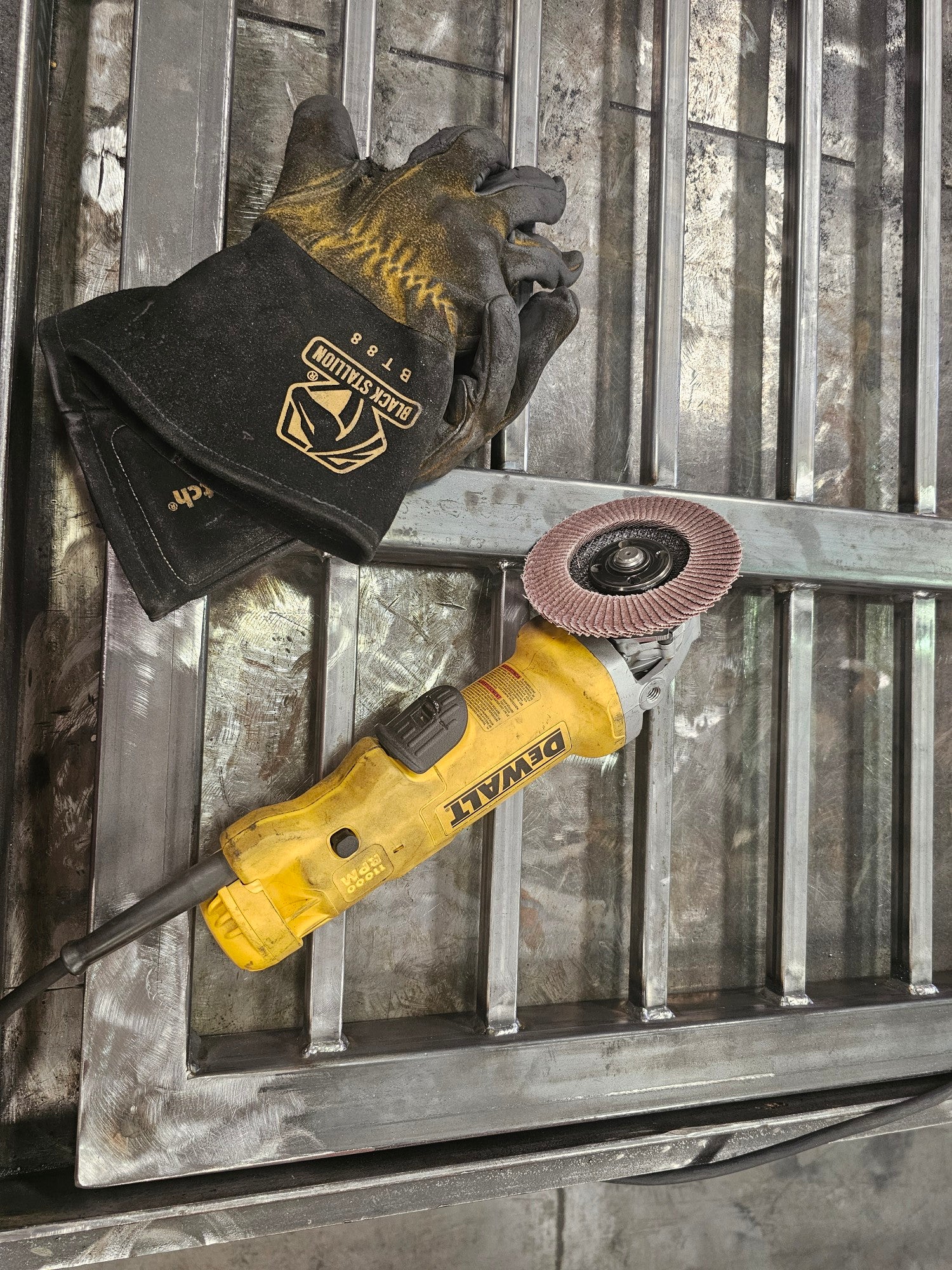 Yellow DeWalt angle grinder and black work gloves resting on an unfinished metal gate frame in a workshop.