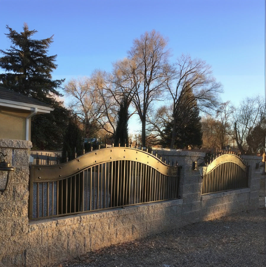 low sun highlighting bronze color arched fence panels with vertical bars and wide metal band at top with rivets and spear finials set in stone block wall surrounding house