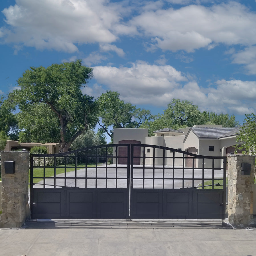 black iron arched double gate with solid paneled botton and grid of squares on top between stone columns in front of stucco chateaux style home. Blue sky, white clouds and green trees in background