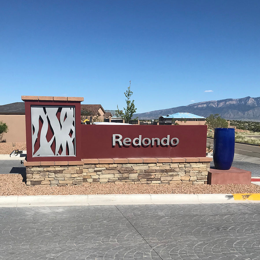 rust color metal sign that says Redondo and stainless steel metal cutout of abstract tree trunks built on stack stone pediment in front of entrance to neighborhood, mountains in backround, southwestern setting