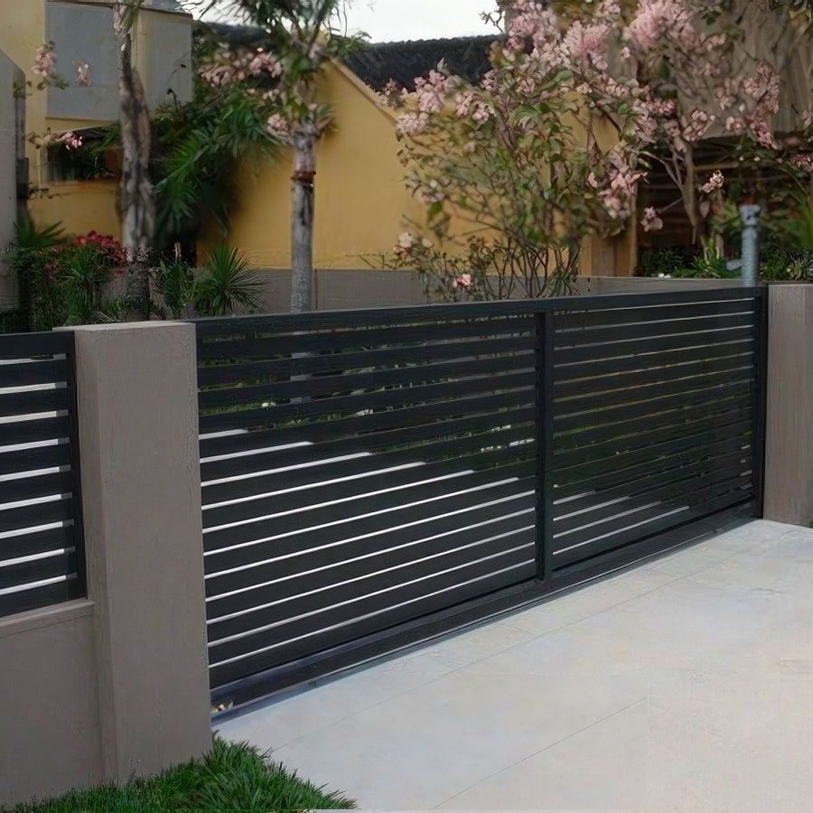 modern horizontal dark metal gate set in gray adobe wall with matching fence panel on driveway and palm trees behind