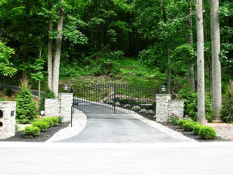 Black wrought iron gate with a curved design, set between stone pillars, leading up a paved driveway by lush greenery.
