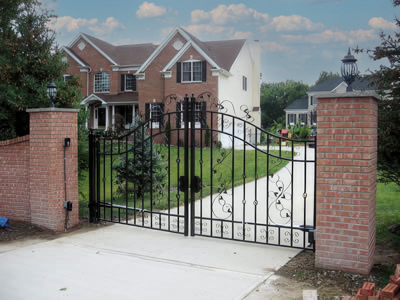 Black iron gate with scrollwork and curved top between brick pillars, leading to a red brick house with a paved driveway.