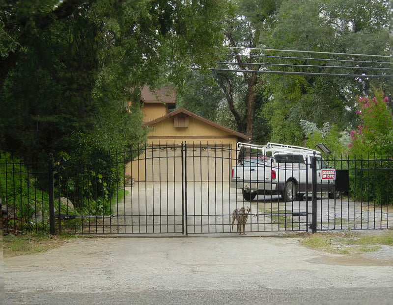 Black metal driveway gate with vertical bars, leading to a tan house with a truck parked in the driveway and trees around.